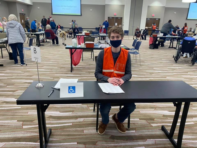 A man wearing a black face mask and orange vest is seated behind a brown table. Behind him are several more tables spaced out through a large banquet hall. There are several people either walking around or sitting in chairs. Photo from Champaign-Urbana Public Health District Facebook page.
