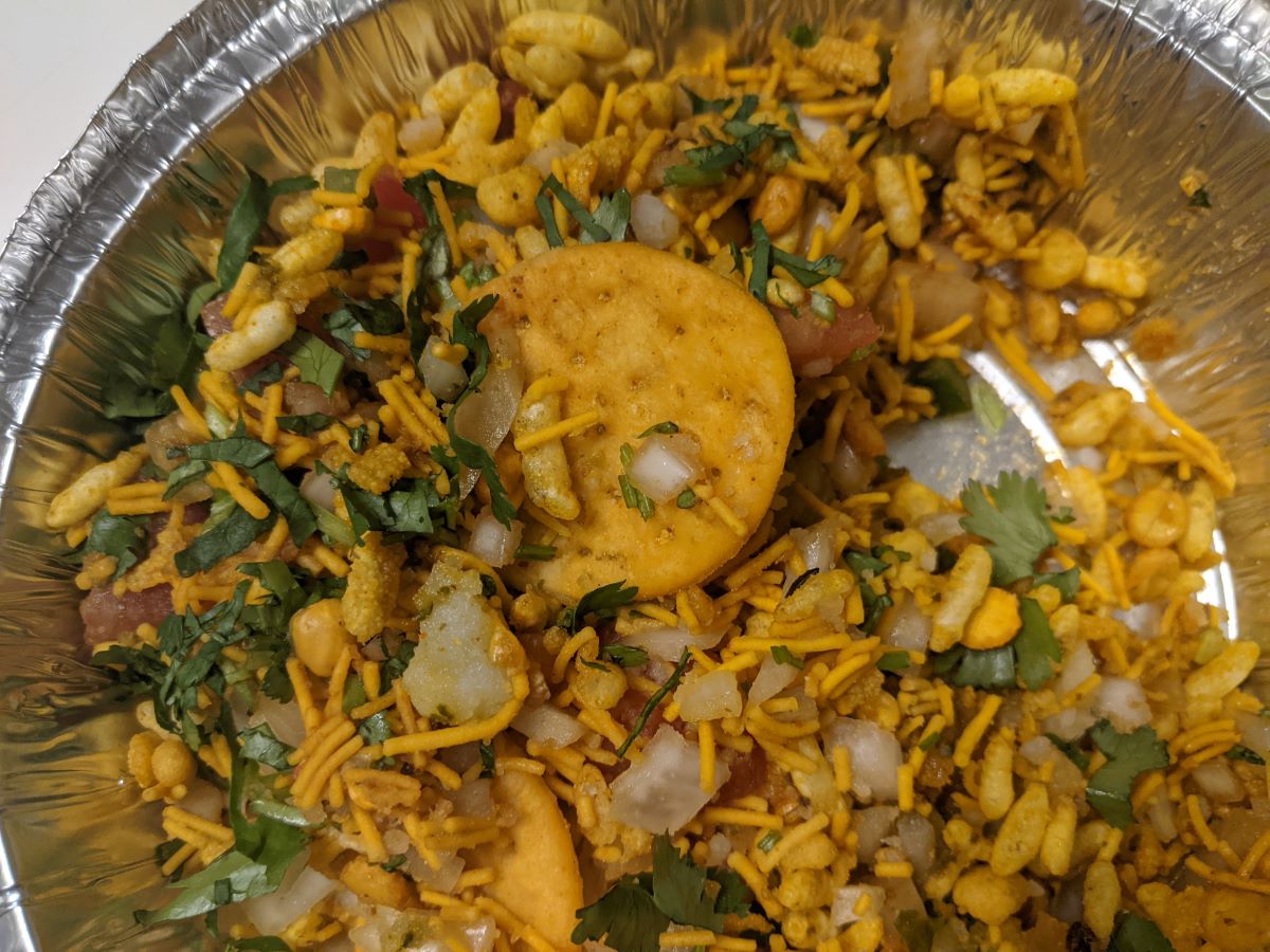 A yellow cracker-like puri is embedded in the bhel mixture.  It is surrounded by yellow noodles, green cilantro, and white chopped onions.  The dish is served in a metal container. Photo by Tias Paul.