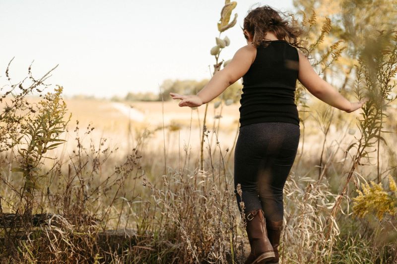 A young girl is facing away from the camera, walking through prairie grass with her arms outstretched. She is wearing a black sleeveless shirt, dark gray leggings, and brown cowboy boots. Photo by Anna Longworth.