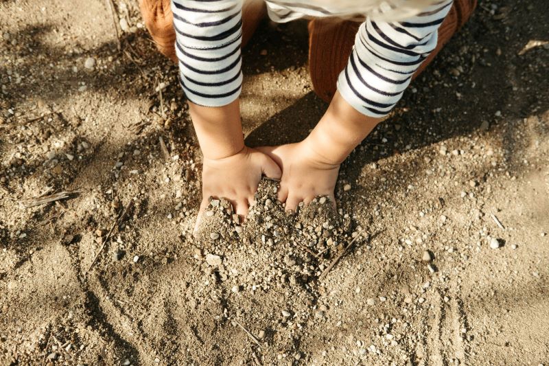 A child is kneeling in the dirt, digging their hands into it. They have a white shirt with black stripes. Photo by Anna Longworth.
