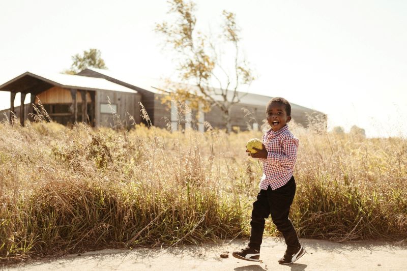 A child wearing a red checked shirt and jeans is holding a yellow ball. He is standing in front of a field of tall prairie grass. A large building with wood siding and a white roof is in the background. Photo by Anna Longworth.