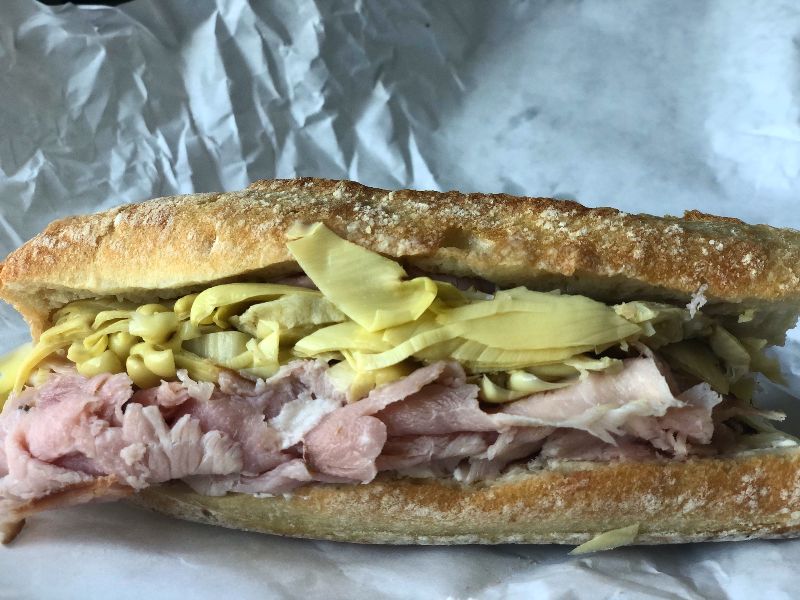 A large baguette sandwich from Cheese & Crackers called France Meets Italy sits on white parchment paper. There are many cold, light green artichokes atop pink shaved ham with a white spread beneath. Photo by Alyssa Buckley.