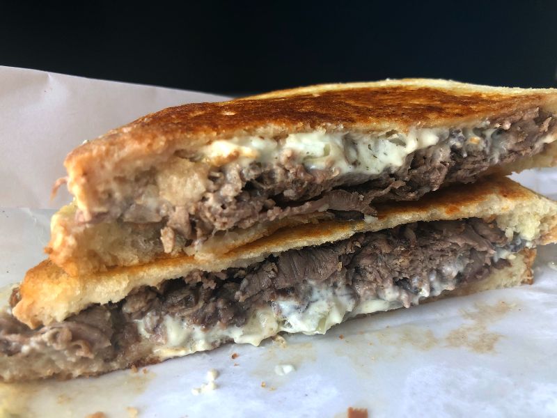 The roast beef on this sandwich is shaved thin and stacked tall inside golden brown sourdough bread. There is a lot of blue cheese spread on the bottom half of the sandwich, oozing a bit out the middle onto the white parchment paper. Photo by Alyssa Buckley.