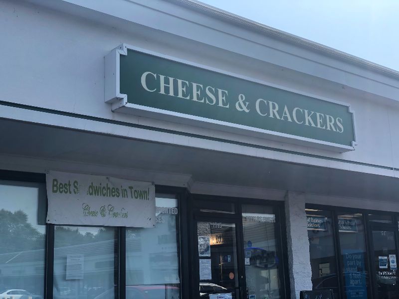The exterior of Cheese & Crackers in Champaign. The sign overhead has the store's name on a white building. Outside, a vinyl banner reads 