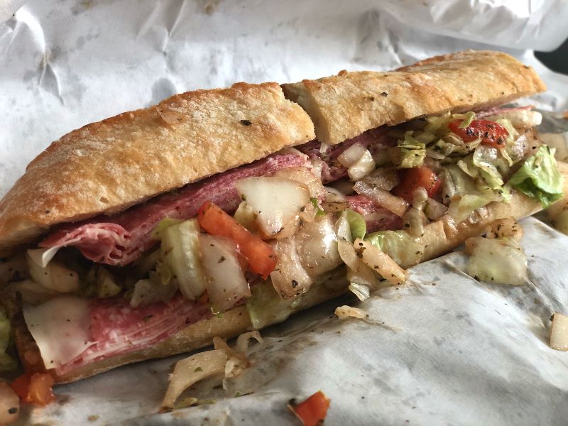 A large Italian sandwich on a baguette from Cheese & Crackers sits on white parchment paper. The salami is plentiful, and the cheese is thin. The veggies are covered in an herby dressing and fall out of the sandwich. Photo by Alyssa Buckley.