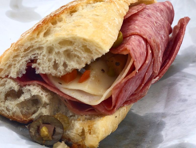 A sideview of a baguette Mortadella sandwich from Cheese & Crackers in Champaign. The bread, sliced cheese, Italian meat, and olive mix are visible from the side. Photo by Alyssa Buckley.