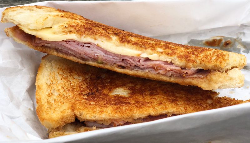 A sliced sandwich on sourdough bread is stacked on top of each other on white paper. The sandwich has lots of pink ham, a few slices of very light yellow Gouda cheese, and the faintest spread of dark brown fig jam on the bottom. Photo by Alyssa Buckley.