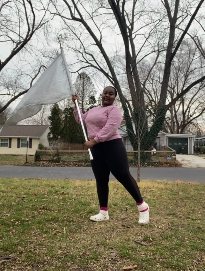A teenage girl who is Black is smiling and posing with a silver flag outside on a cold, cloudy day in a neighborhood. She is holding a flag for marching band color guard. The girl is wearing a pink sweater, black leggings, pink socks, and white crocs. Photo provided by Keyarah Johnson.