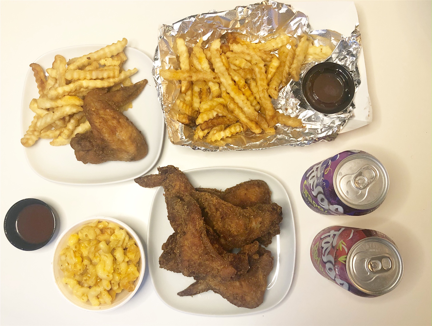 An overhead shot of the author's dinner. On the far left, there is a small square white plate with one wing and fries. Beside it, a basket of crinkle fries and below a white plate of six wings. There is a cup of hot sauce plus a cup of macaroni and two Faygo sodas. Photo by Alyssa Buckley.