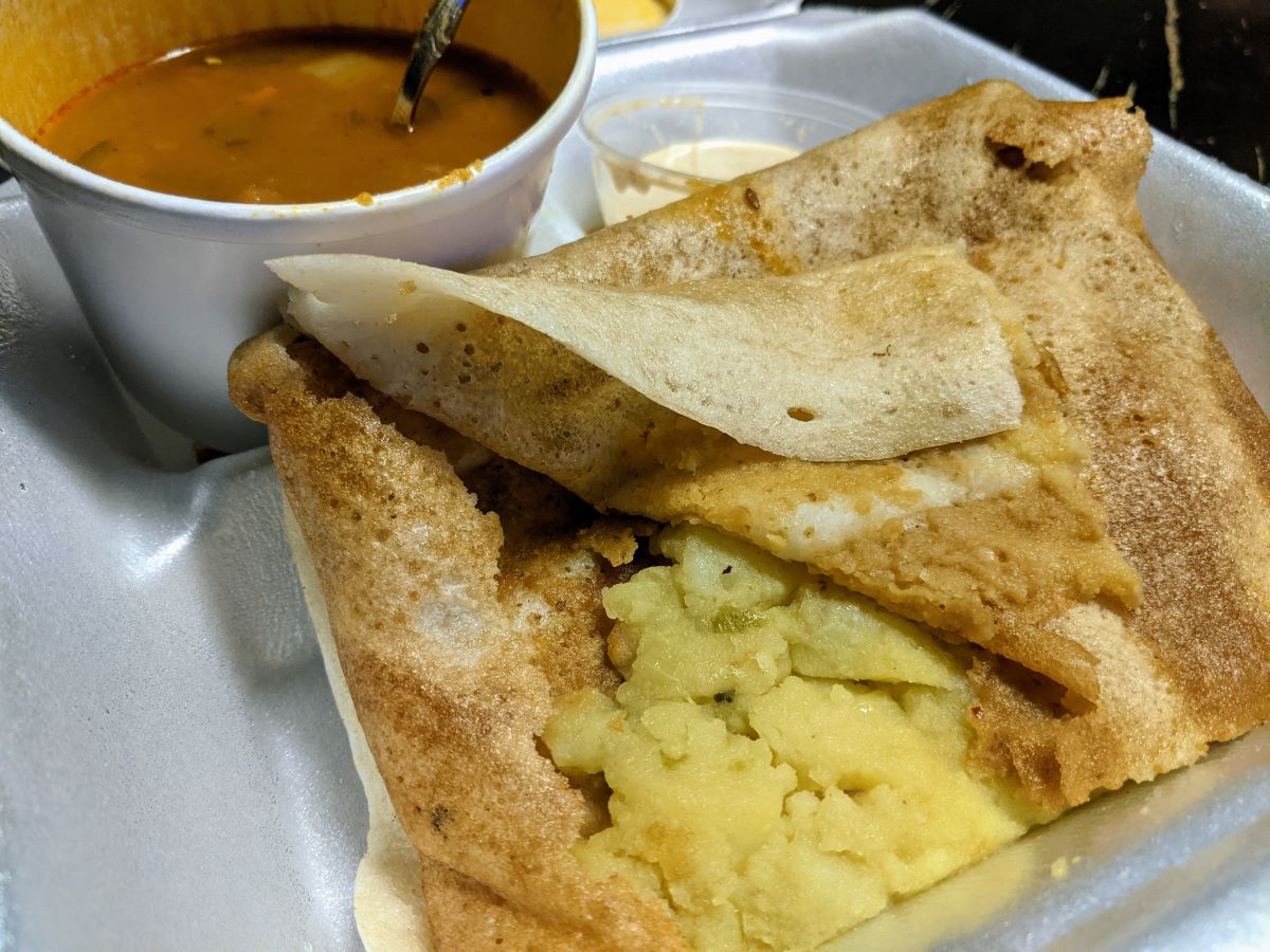 In the picture, we see a crepe stuffed with yellow potato filling. Behind the crepe is a styrofoam cup full of red soup. On the right side of the soup, we can barely see a pale orange coconut chutney in a plastic sealable cup. Photo by Tias Paul.
