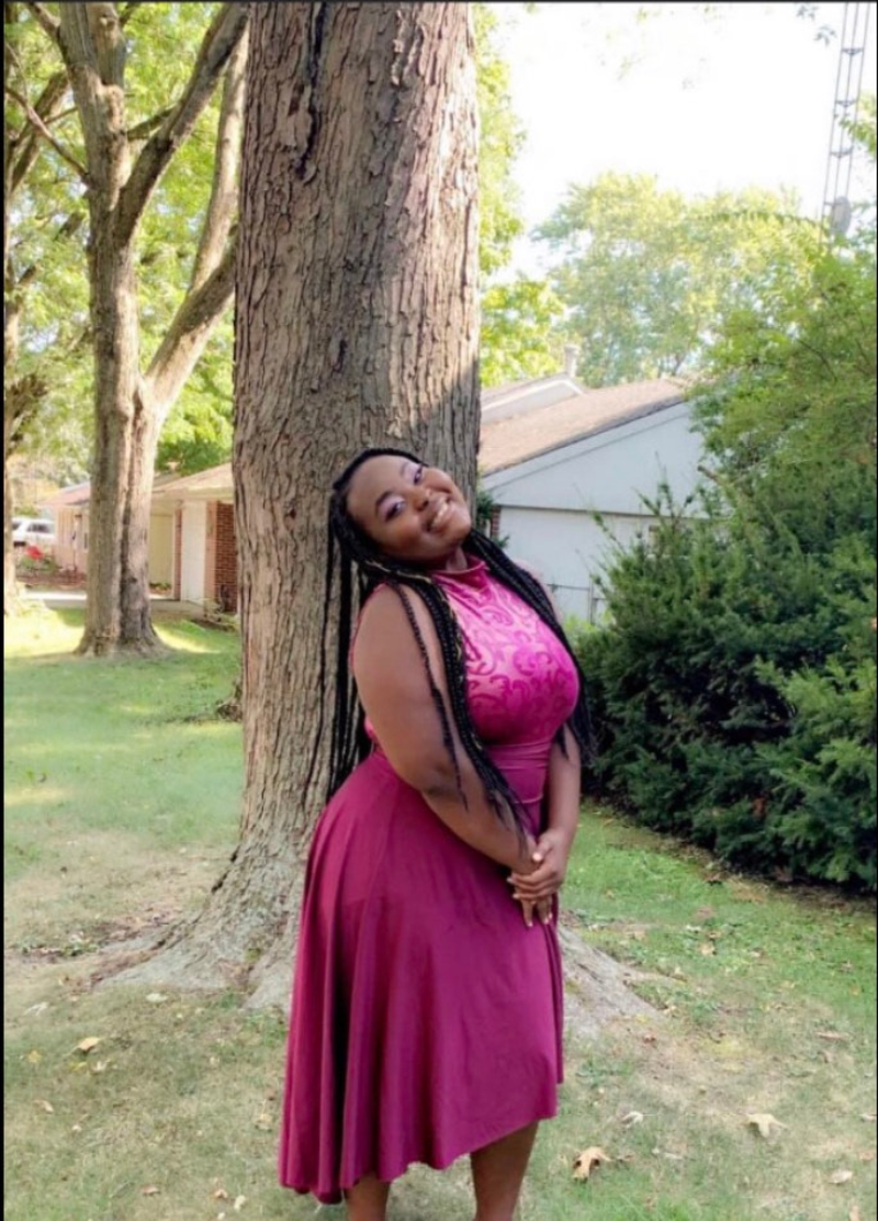 A teenage girl who is Black is wearing a formal knee-length dark pink satin dress, smiling and posing in a yard next to a tree. Her head is tilted back, and she has purple eyeshadow and long box braids. Photo by Keyarah Johnson