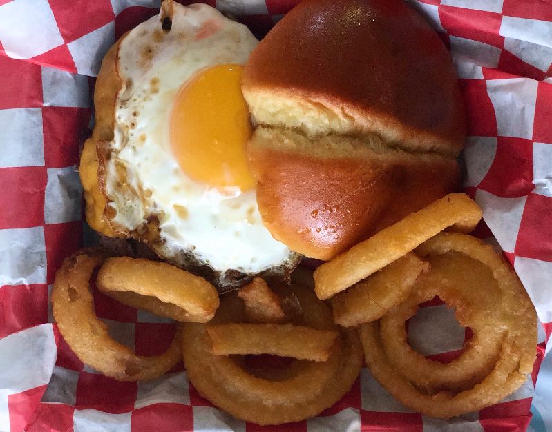 An overhead shot of NAYA's hangover burger which features a sunny side up egg on a burger with a soft bun top beside it. Next to the burger is a side of onion rings in a red-and-white checkered paper underneath. Photo by Alyssa Buckley.