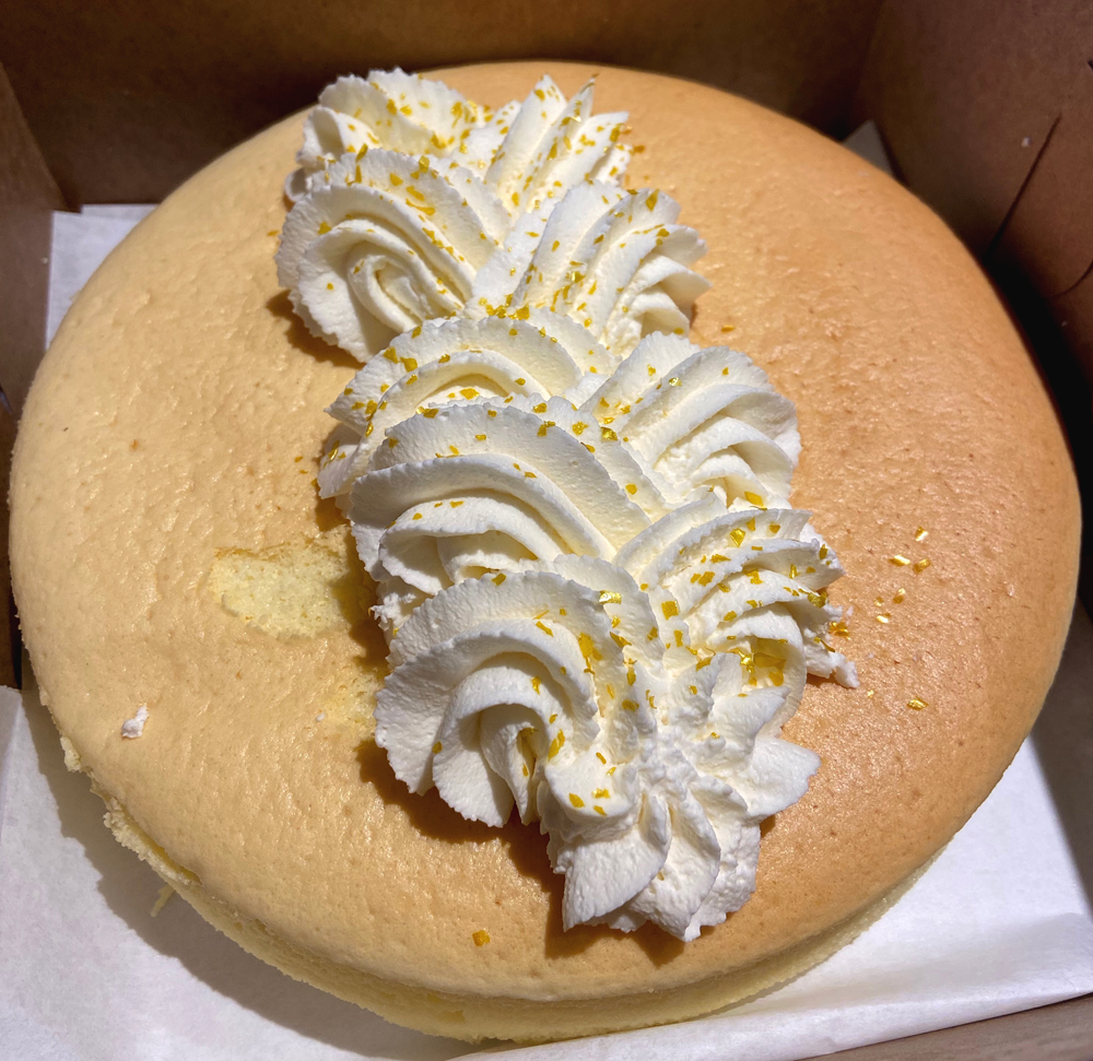 Eight-inch soufflÃ© cheesecake from Suzu's Bakery in Champaign. A lightly golden colored cheesecake sits in a box. The top is decorated wiht a whipped cream braid, dusted with lemon zest, down the center. Photo by Jessica Hammie. 