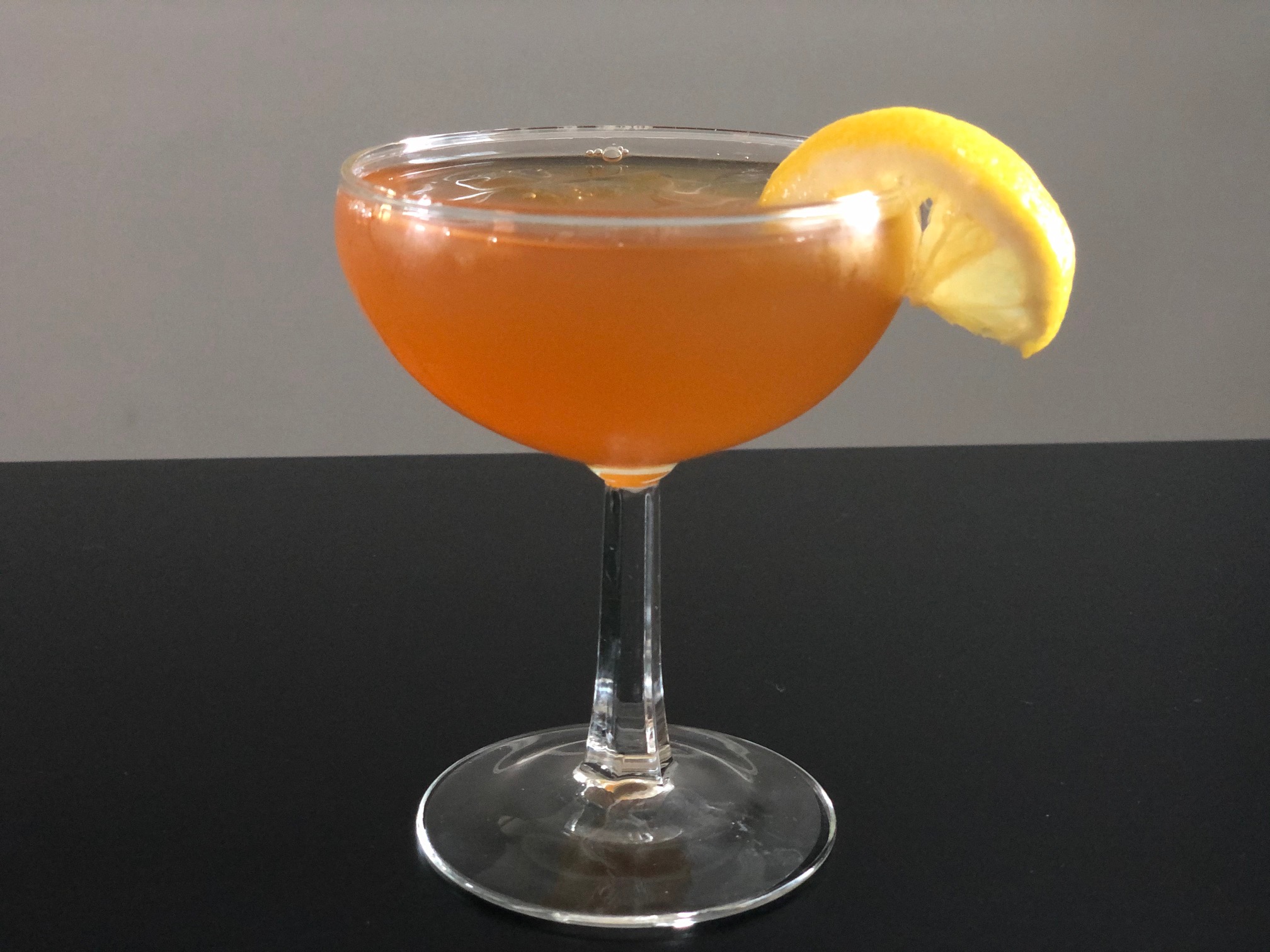 An orange cocktail in a coupe glass on a black table has a lemon garnish. Photo by Alyssa Buckley.