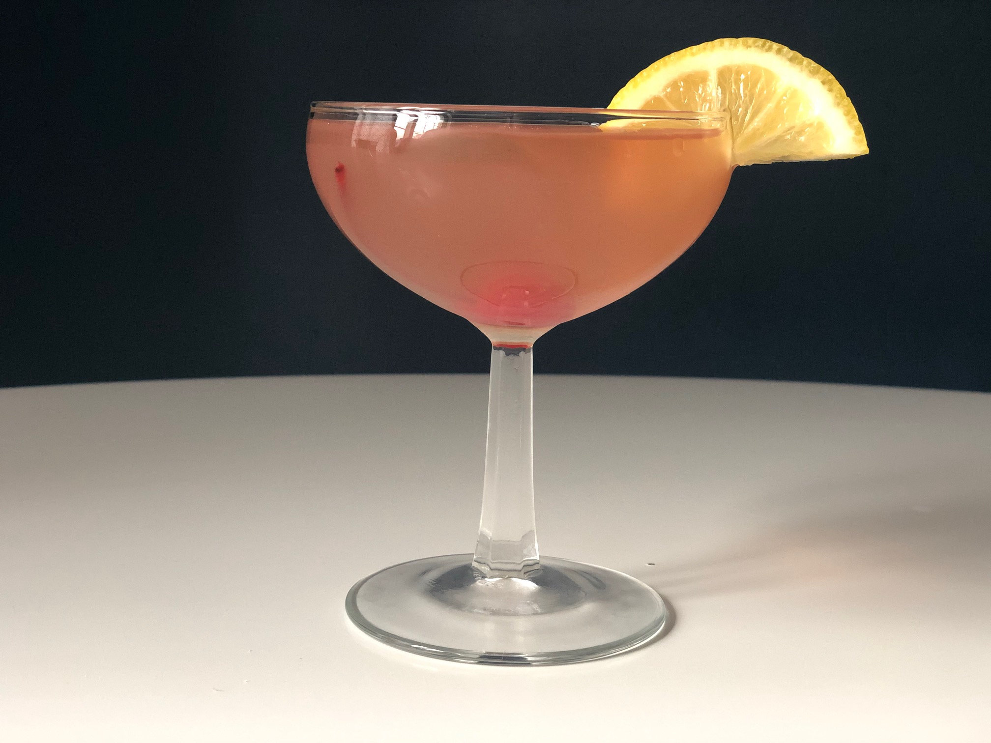 A light red colored cocktail is in a coupe glass with a lemon garnish on a white table. Photo by Alyssa Buckley.