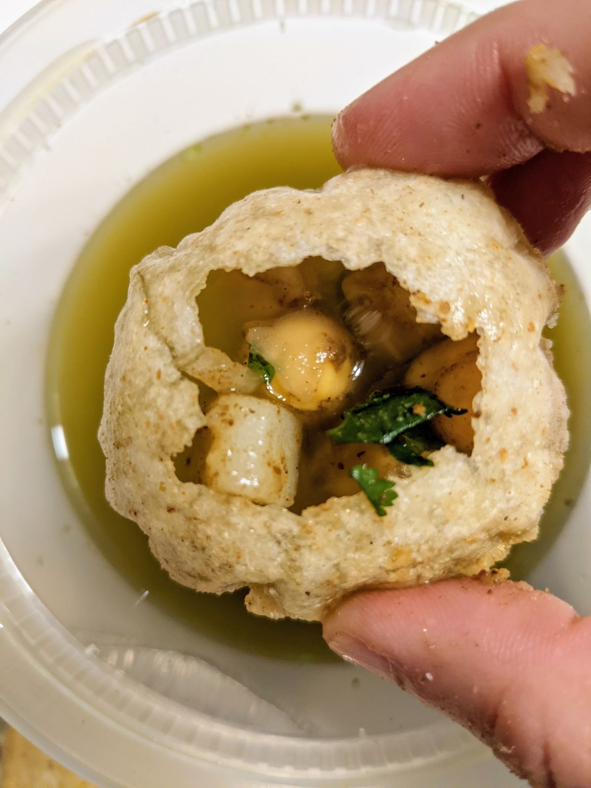 Fingers holding up a crisp round puri filled with chickpea and potato filling and tamarind water.  Underneath the puri, is the container of green tamarind water. Photo by Tias Paul.