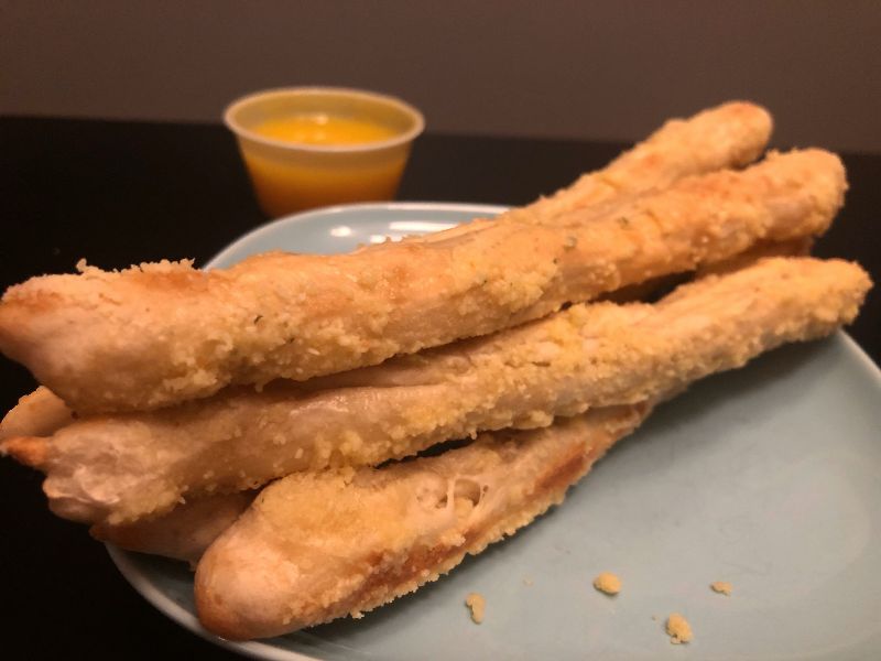 Six long, skinny breadsticks from Po' Boys sit on a white plate on a black table. Behind the plate of breadsticks is a small, plastic cup of garlic butter. Photo by Alyssa Buckley.