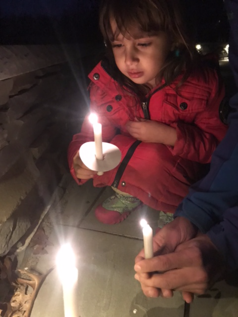 A young girl with dark brown hair and brown eyes is holding a lit candle and looking at the flame. She is wearing a red coat and gray boots, and sheâ€™s squatting on a sidewalk. An adult is sitting next to her, also holding a lit candle, and they are demonstrating how to hold the candle safely. They are outside at night time. Photo by Darya Shahgheibi. 
