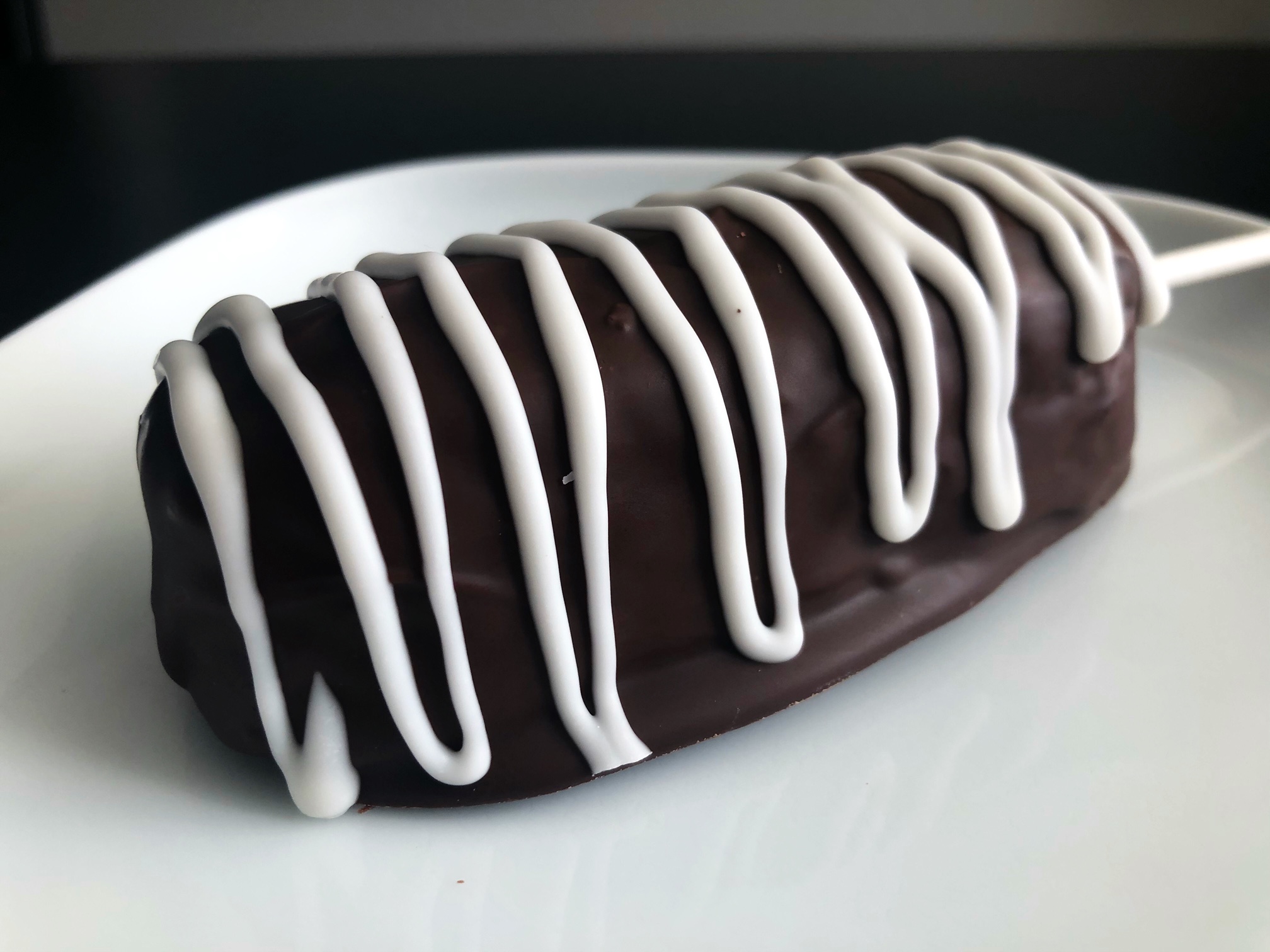 A chocolate covered Twinkie is drizzled with white chocolate and laying on a white plate. There is a white stick for easy holding. Photo by Alyssa Buckley.
