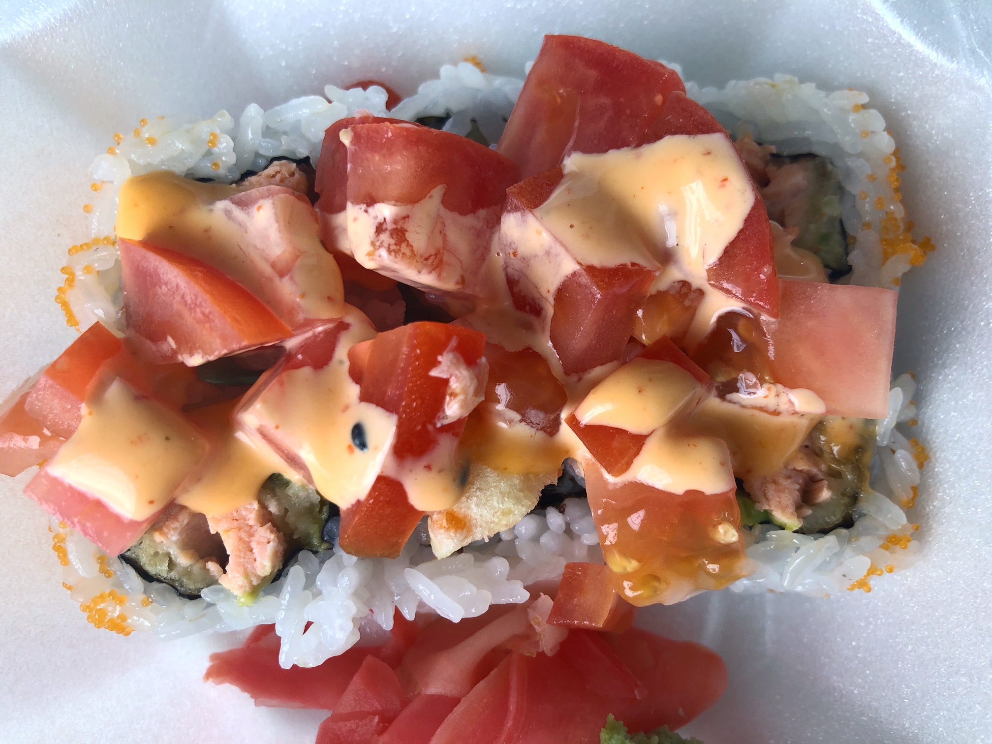 In a white styrofoam container, there is an Alaska Roll from Sushi Kame. The sushi roll is topped with a lot of diced tomato plus a generous drizzle of an orange sauce. Photo by Alyssa Buckley.