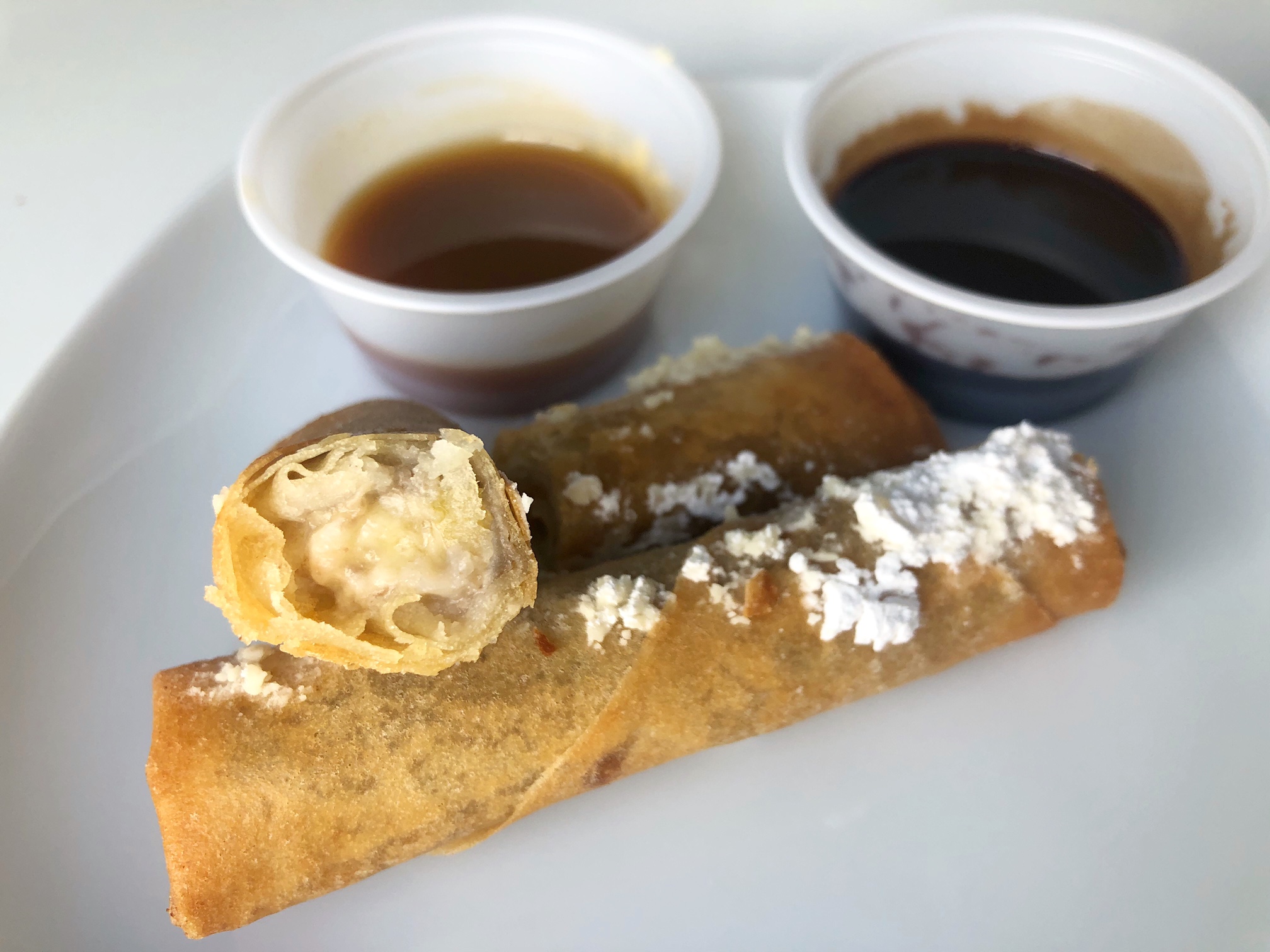 Two dessert egg rolls are on a plate, one sliced open to show the banana filling. Behind the white plate are two dipping sauces: caramel and chocolate. Photo by Alyssa Buckley.