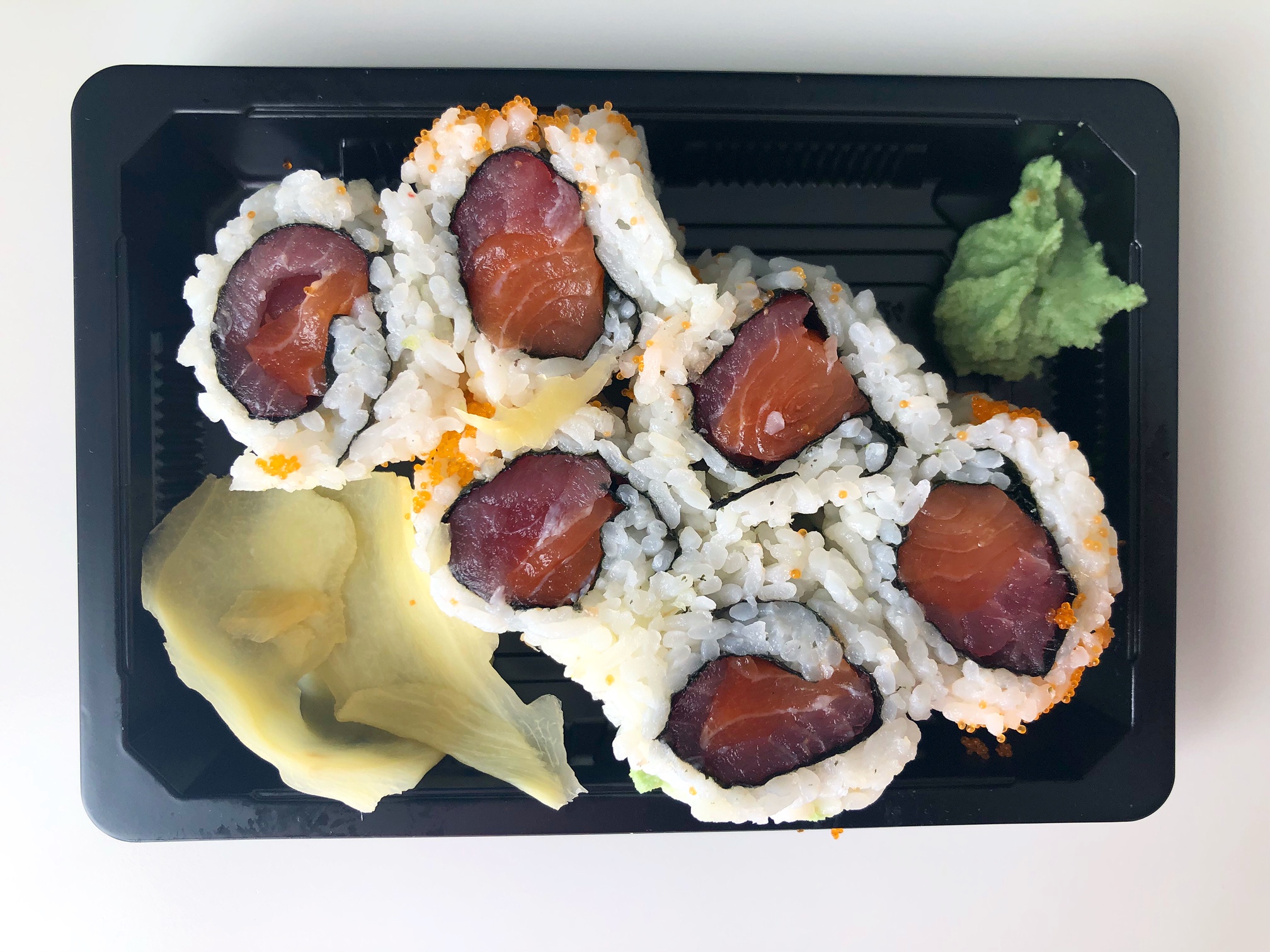 An overhead photo of the New York roll from Sushi Ichiban in a black plastic takeout container with ginger and wasabi. The roll has two pink fish wrapped inside seaweed with white rice and orange fish eggs outside. Photo by Alyssa Buckley.