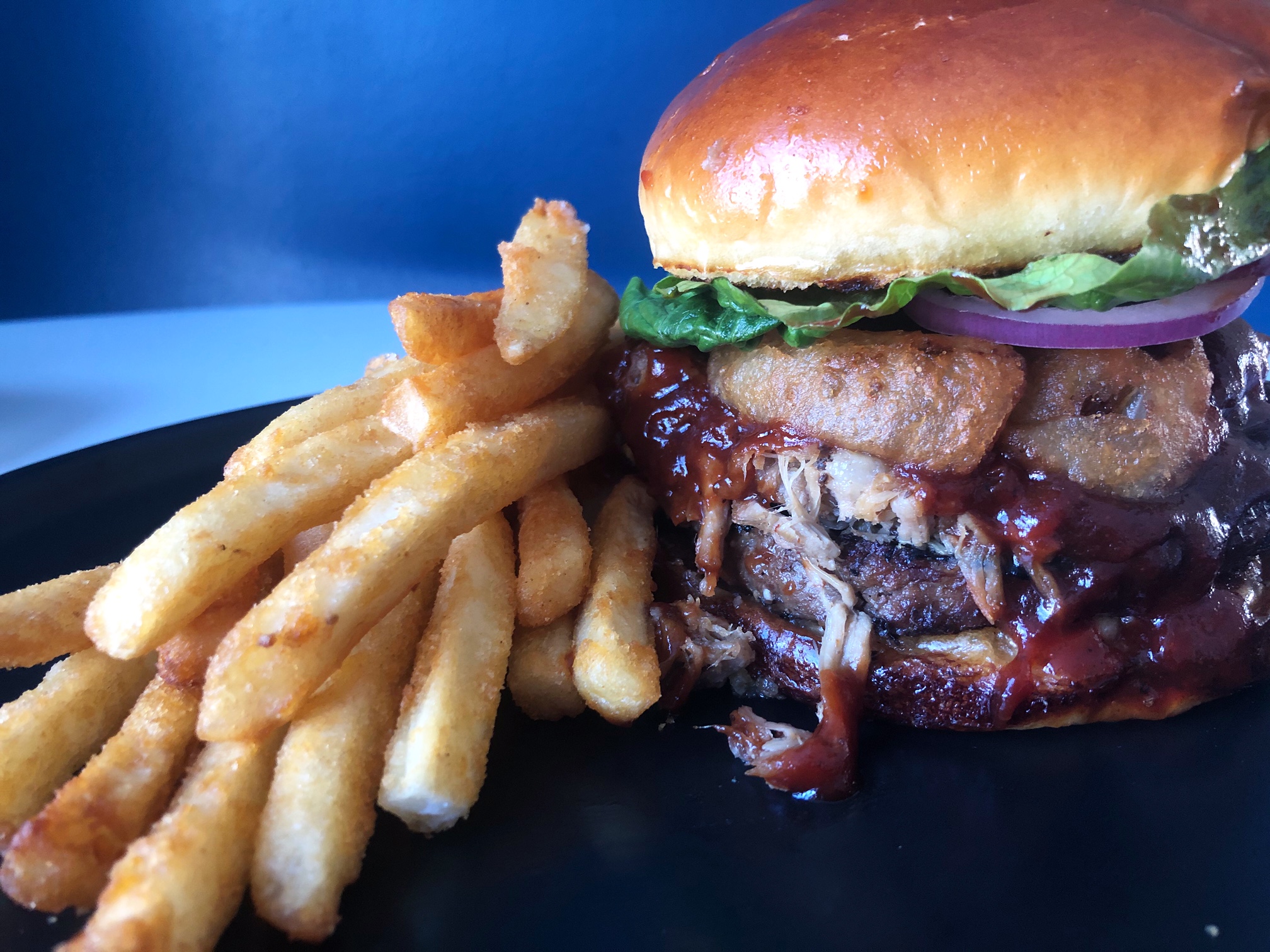 A burger with bbq pulled pork and onion rings sits on a black plate besides a lot of french fries. Photo by Alyssa Buckley.