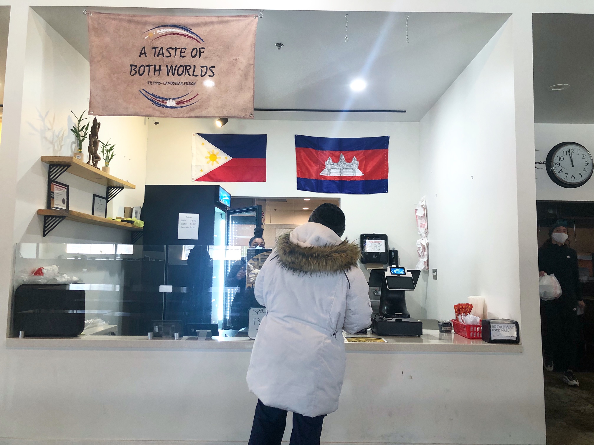 The newest vendor at Broadway Food Hall is A Taste of Both Worlds. A flag with the name of the restaurant is in the top corner of an all white ordering front of house. There is a woman in a white puffer coat ordering. Photo by Alyssa Buckley.
