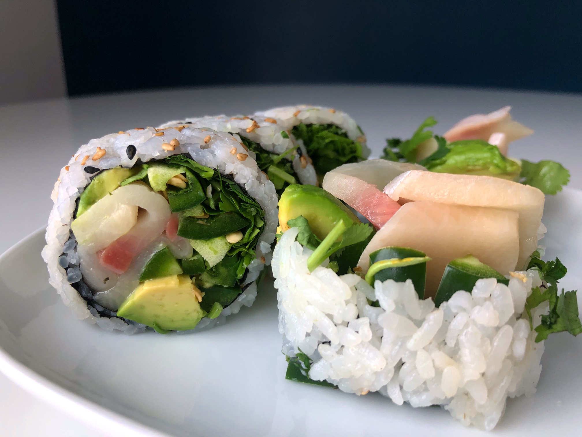 This Mexican roll has yellowtail, cucumber, and large bunches of cilantro wrapped in white sushi rice. The roll is on a white plate with a navy wall behind it. Photo by Alyssa Buckley.