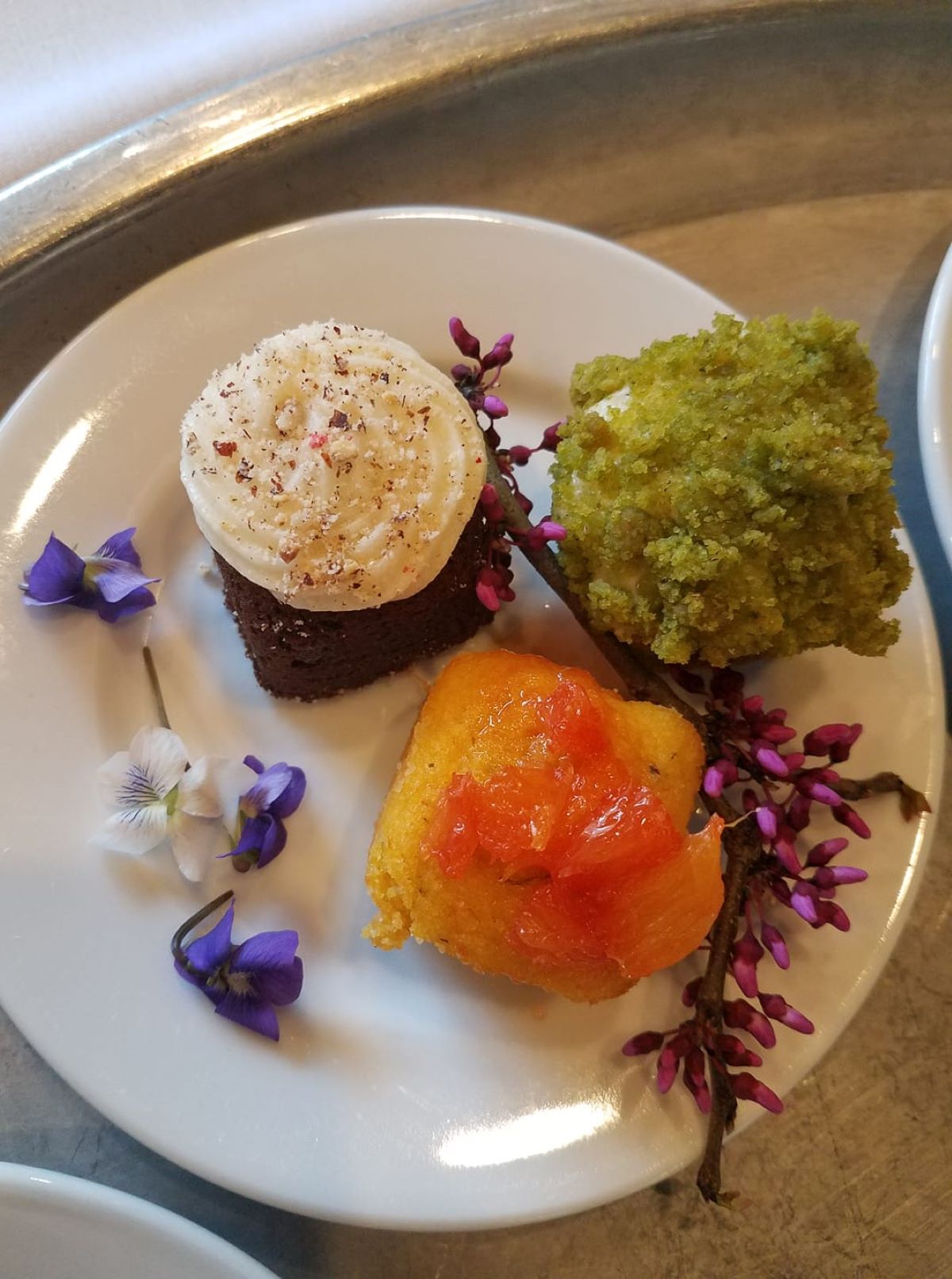 From Allertonâ€™s Forest to mansion dinner, in 2018, three desserts, brown/white, orange, and green, on a round, white plate. The plate is decorated with purple and white violets and redbud twig with closed buds. Photo by Nate Becca.