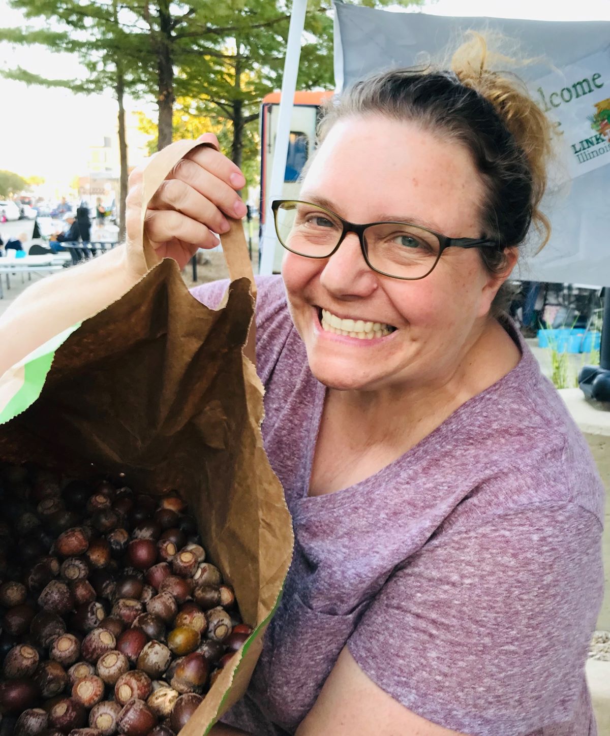 Heidi is wearing a purple shirt and black glasses.  She is holding open a brown paper bag of acorns. Photo by Eliana Brown.
