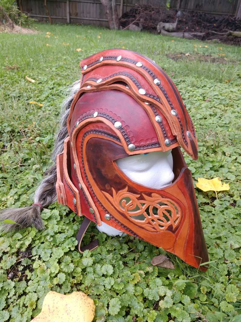 A finely worked leather helmet rests on a mannequin head in a field of grass.