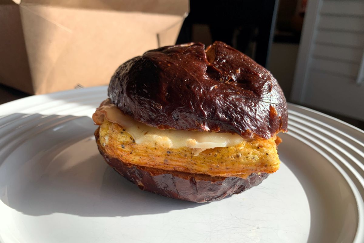 A side view photo of a breakfast sandwich on a white plate. The sandwich is a dark brown pretzel bun with melted cheese on top of a square piece of scrambled egg. Photo by Megan Friend.