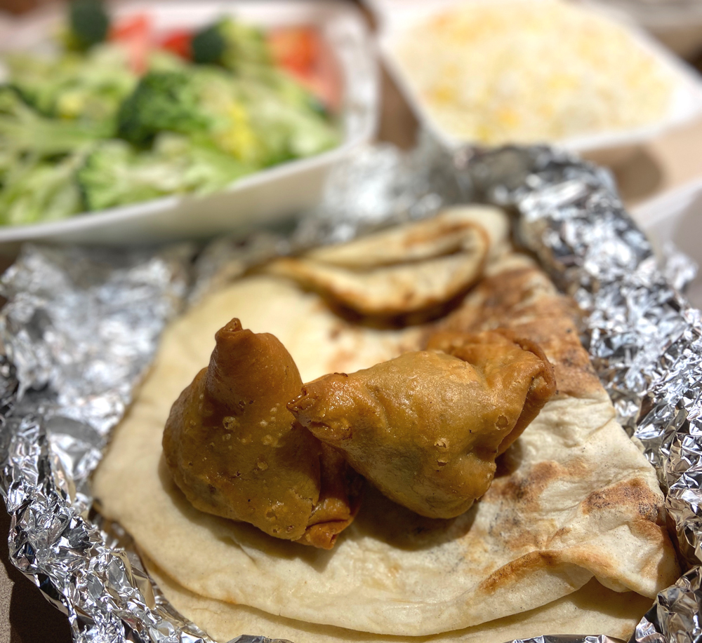 Two samosas sit on top of naan, which are all on top of tin foil. Food from Kohinoor. The samosas are pyramidal shaped fried food. Photo by Jessica Hammie. 