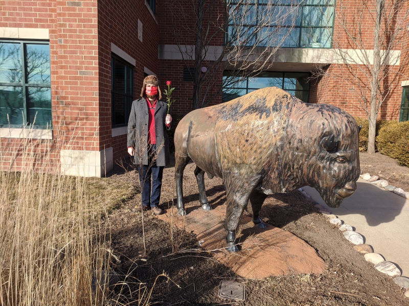 The writer is standing next to a statue of a buffalo. He has a winter hat with flaps, a red mask, and is holding a single red rose. Photo by Andrea Black.
