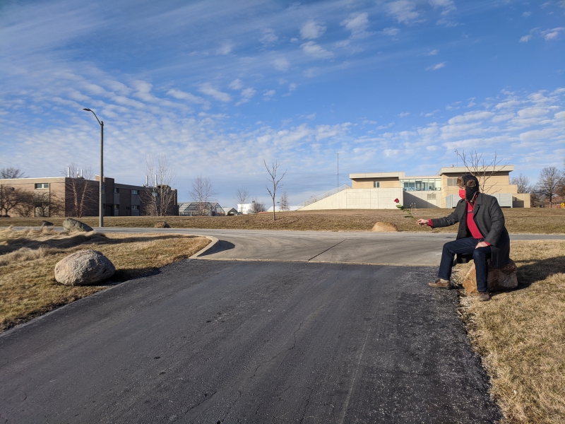 The writer is sitting on a boulder next to a road, arm outstretched, holding a red rose. There is a similar boulder on the opposite side of the road. Photo by Andrea Black. 
