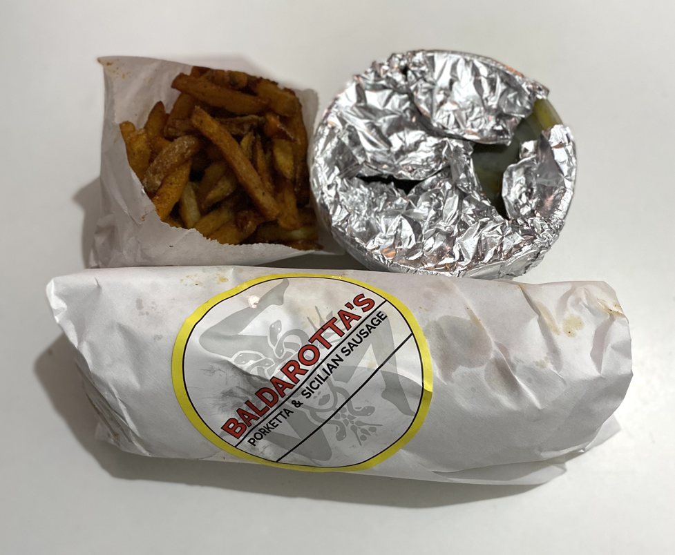 Baldarotta's Wednesday special. A white parchment paper wrapped sandwich with a sticker of Baldarotta's logo affixed to it sits on a white table below a side of fries and a tin-foil wrapped circle side. Photo by Patrick Singer.