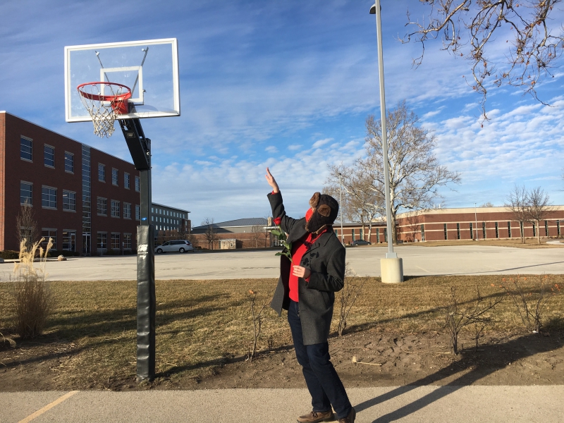 The writer is standing on a concrete basketball court, just adjacent to a basketball hoop with a clear backboard. He is gesturing like he's shooting a basket with one hand. Photo by Andrea Black.