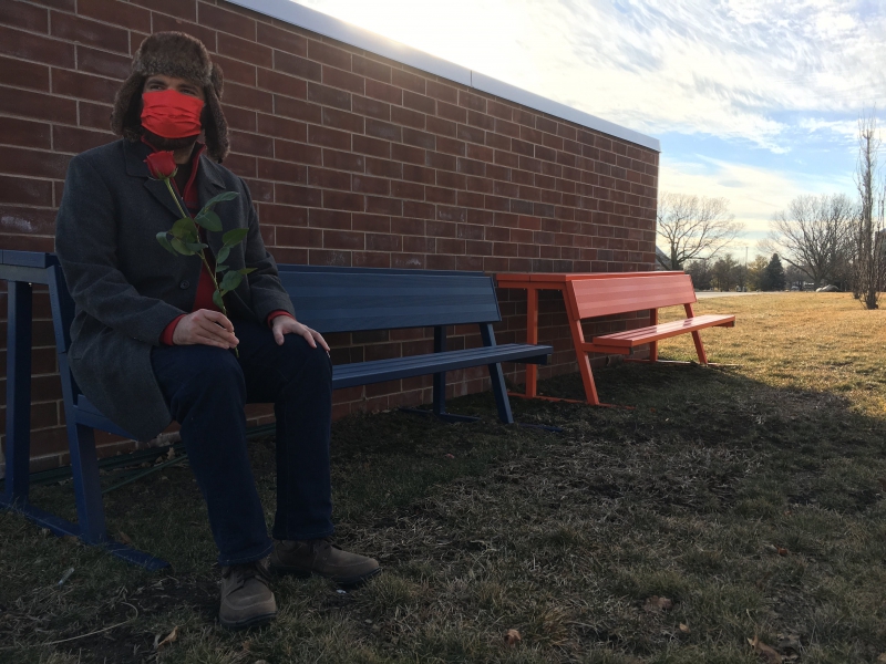 The writer is sitting on a blue metal bench. There is an orange metal bench next to it. The benches are sitting along a brick wall. Photo by Andrea Black.
