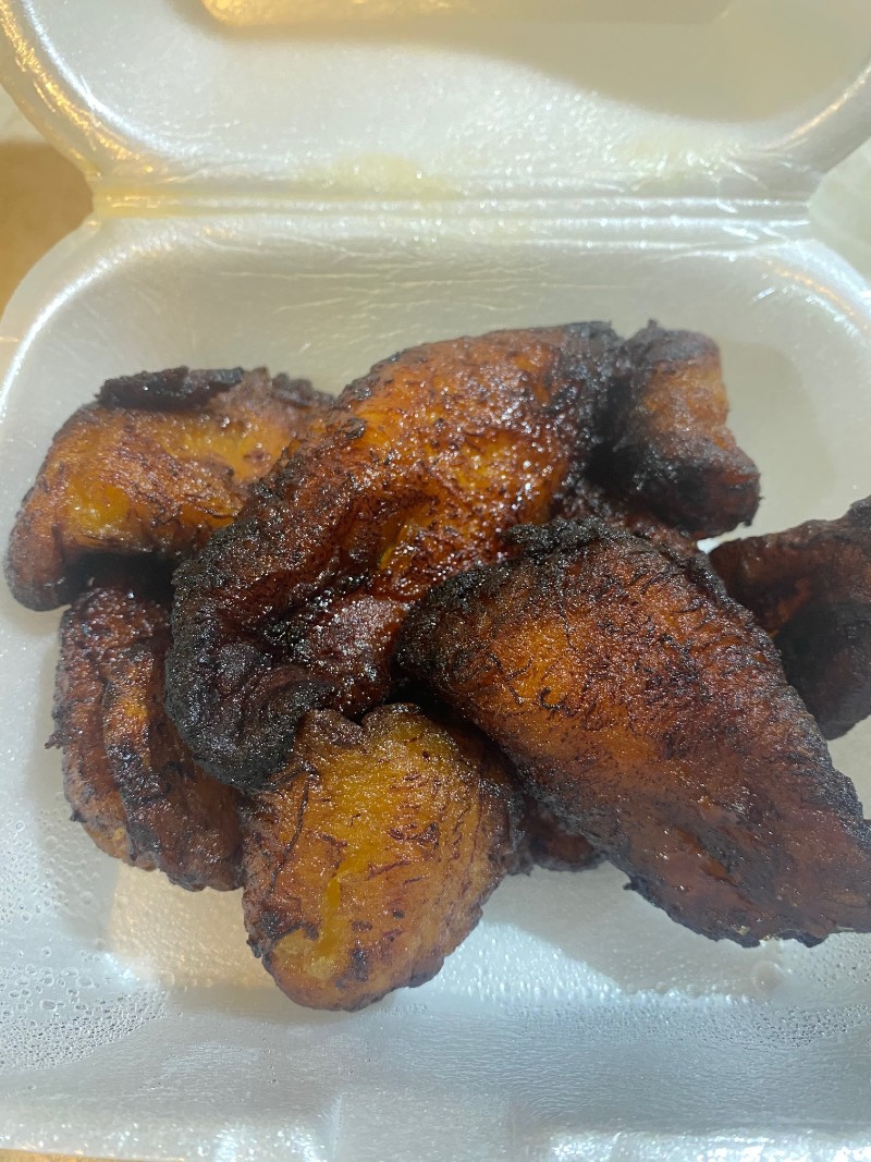 A cluster of cooked plantain pieces are in a square, white Styrofoam container. They are yellow with some brown coloring from caramelizing with blackened edges. Photo by Julie McClure.
