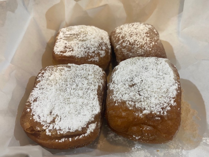 Four golden brown rectangular beignets are in a white Styrofoam container. They are coated with white powdered sugar. Photo by Julie McClure.