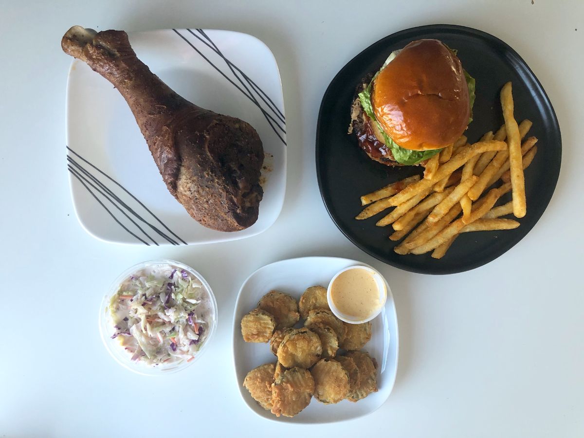 An overhead photo of a dinner from Neil St. Blues in Champaign. There is a turkey leg on a white plate, a burger and fries on a black plate, a cup of coleslaw, and a small plate of fried pickles all on a white table. Photo by Alyssa Buckley.