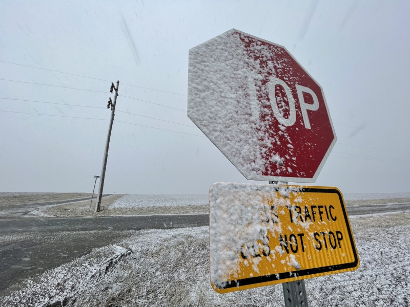 A stop sign is partially covered with a layer of snow. Behind it is a crossroads, with fields on either side. Photo by Andrew Pritchard.