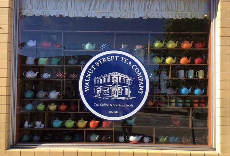 Walnut Street Tea Company window with their logo on the side that says Tea, Coffee, and Specialty Foods. A rainbow of tea kettles are seen behind the window logo. Photo provided by Walnut Street Tea Company.