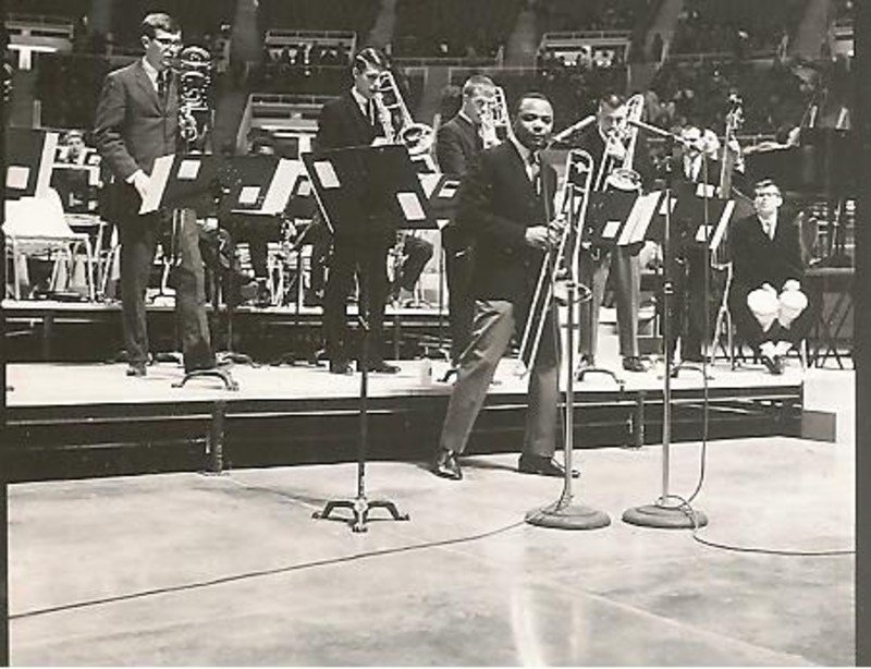 A black and white photo. A man in a suit is holding a trombone, and standing in front of a small stage. There are several other musicians on the stage. In the foreground there are a few microphones on stands. Photo from Champaign Urbana History Facebook page.