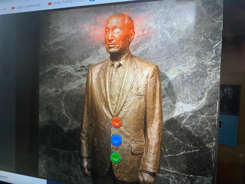 A screenshot of a statue of Arnold Beckman. Red, blue, and green buttons have been superimposed over the image. There is a red light emanating from his left eye. Screenshot by Julie McClure.
