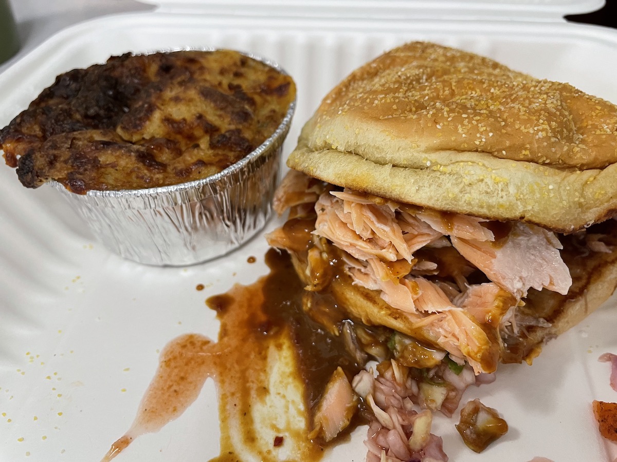 A pulled salmon sandwich from Black Dog sits on a white plate beside a tin foil cup of twice baked potato casserole. Photo by Anthony Erlinger.