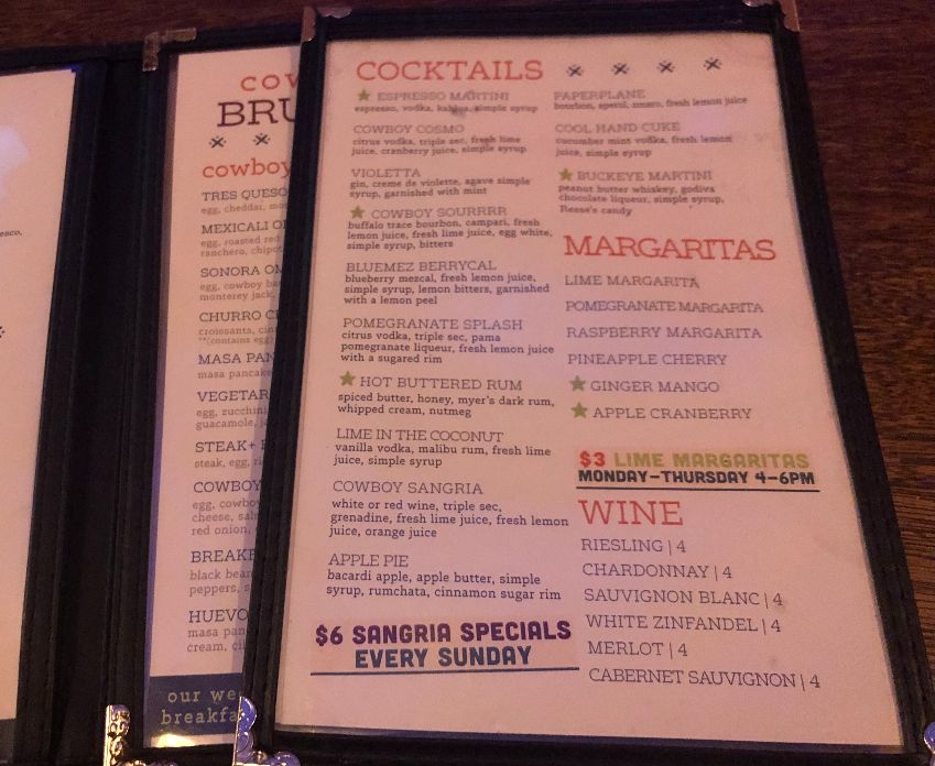 The drink menu from Cowboy Monkey sits atop the food menu on a wooden bar in low lighting one evening in Downtown Champaign. Photo by Alyssa Buckley.