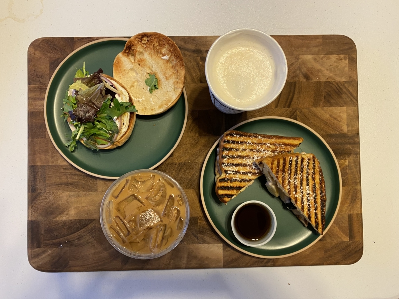 An overhead photo of the author's Cafe Kopi order. On a green plate on the top left, there is a bagel sandwich opened revealing a mixed greens topping. Beside it on the top right, there is an open cup of a latte. Below it on a green plate, there is a diagonally cut sandwich with a small side cup of a dark liquid. Beside it on the bottom left, there is an iced coffee drink. Photo by Mia Hanneken.
