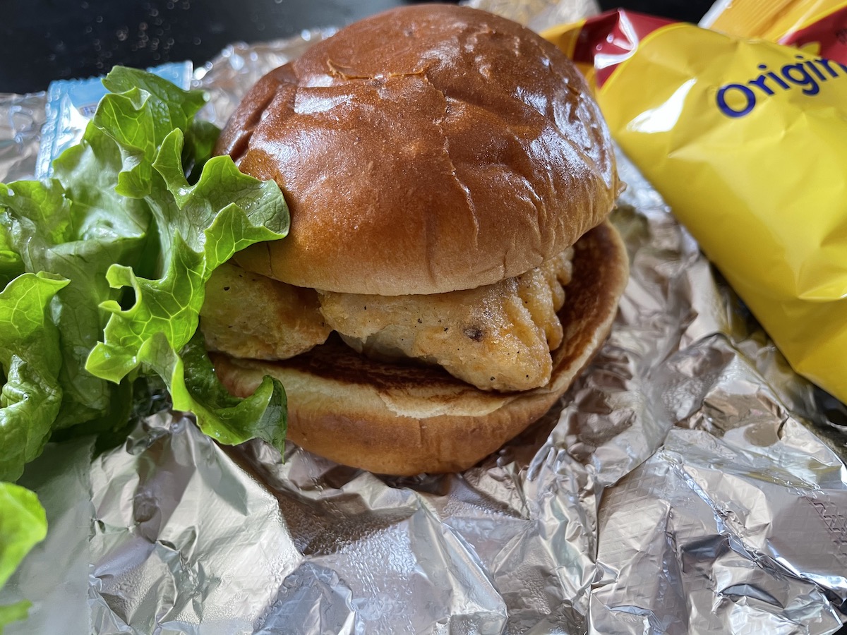 A fried fish sandwich from Esquire Lounge in Champaign sits on a tin foil wrapper with a bag of Lay's potato chips in the background. Photo by Anthony Erlinger.